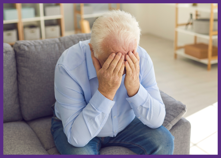 photo of an exhausted senior man sitting on a sofa, holding his hands over his face due to caregiving stress