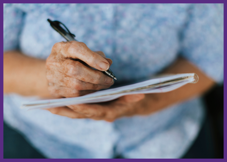 up close photo of a senior woman writing in a notebook she is holding - better manage a chronic disease by taking notes at home and every medical appointment